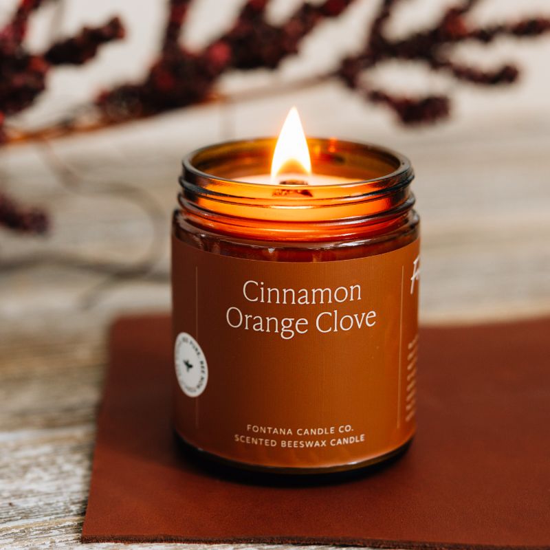 Candle in a jar labeled 'Cinnamon Orange Clove' by Fontana Candle Co. on a wooden surface with blurred background.