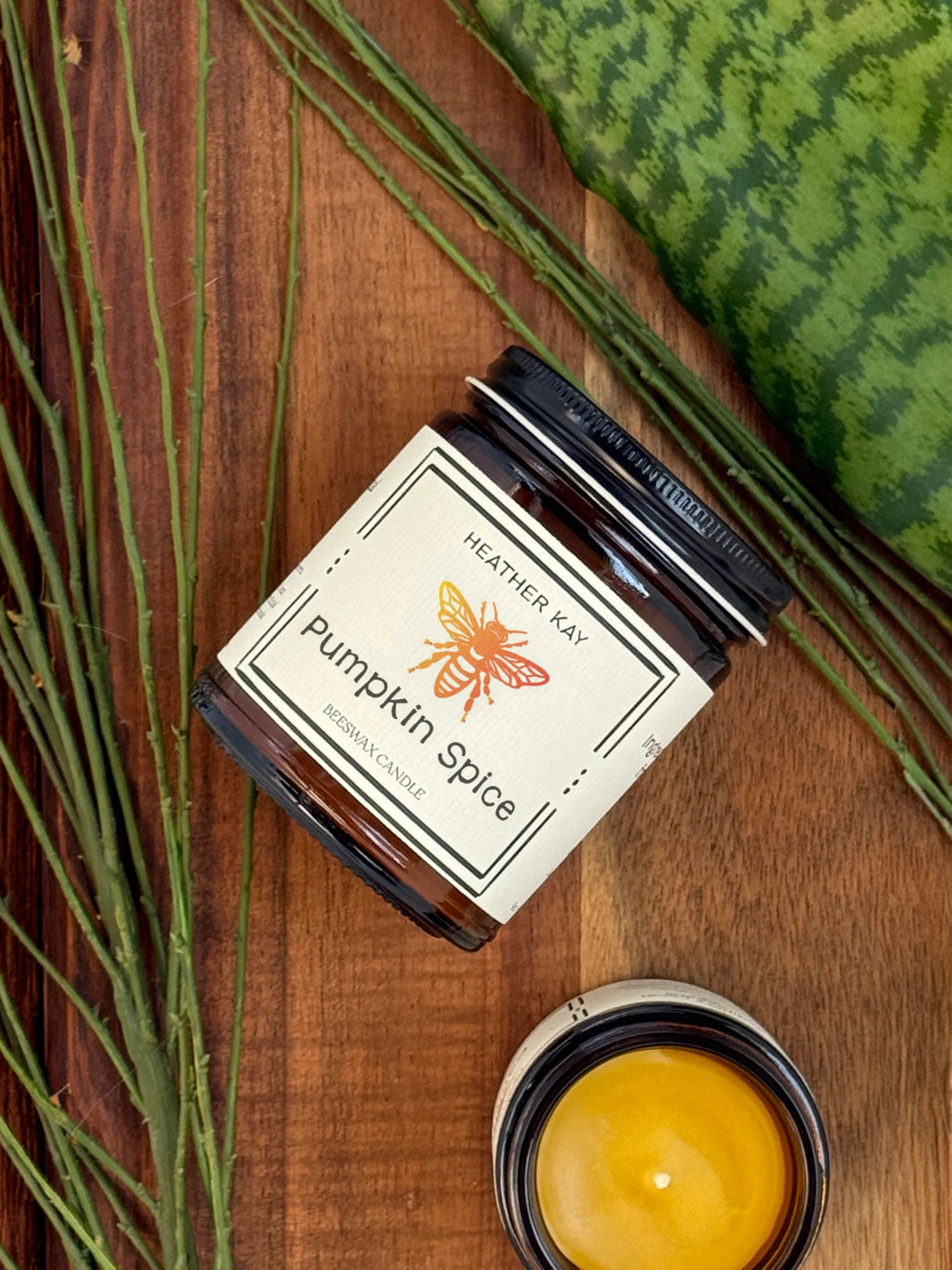 A jar candle labeled 'Pumpkin Spice' by Heather Kay, styled on a wooden surface with green leaves and a watermelon slice. A small open candle jar showing yellow wax is in the foreground.