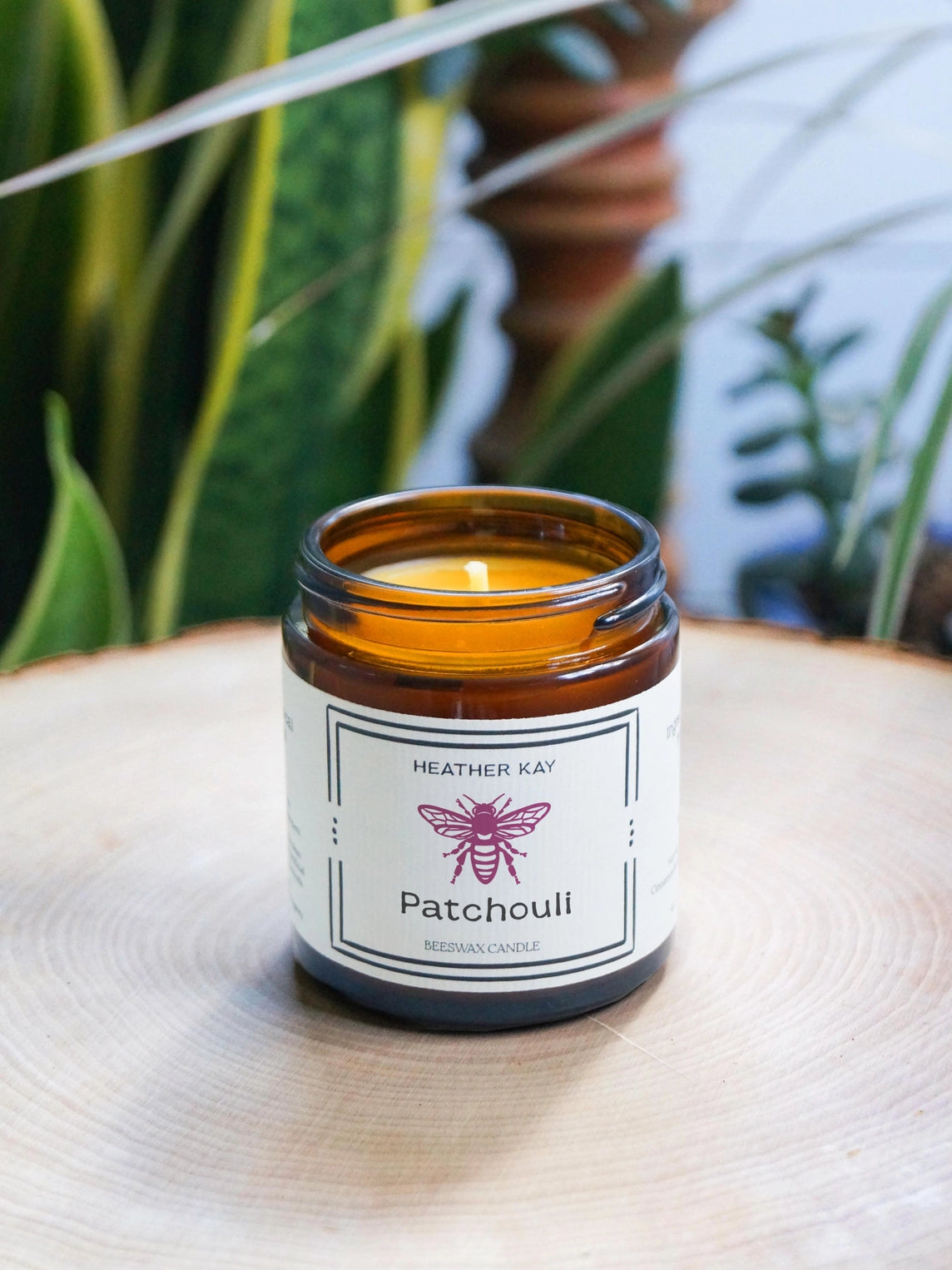 An amber glass jar candle labeled 'Heather Kay Cedarwood Medium Candle' sits on a round wooden slice with blurred green plants in the background.