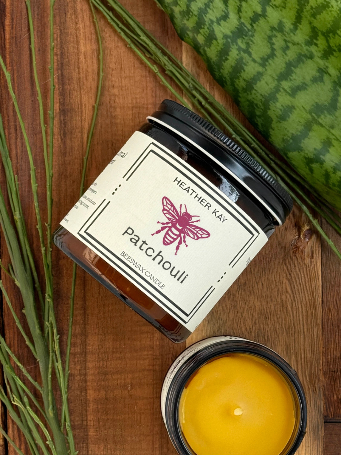 A jar candle labeled Heather Kay Patchouli Brown Wax is styled on a wooden surface with dried green stems, a watermelon slice, and an open candle showing yellow wax.
