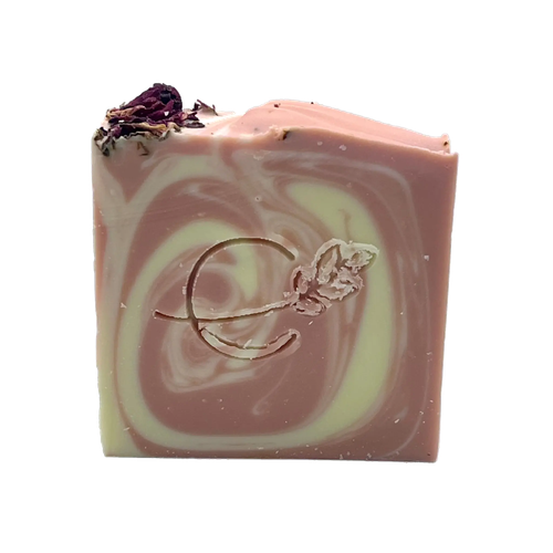 A square bar of handmade soap with a dusty pink and pale yellow marbled swirl pattern, featuring a dried purple flower on top and an embossed rose motif on the front, set against a plain white background.