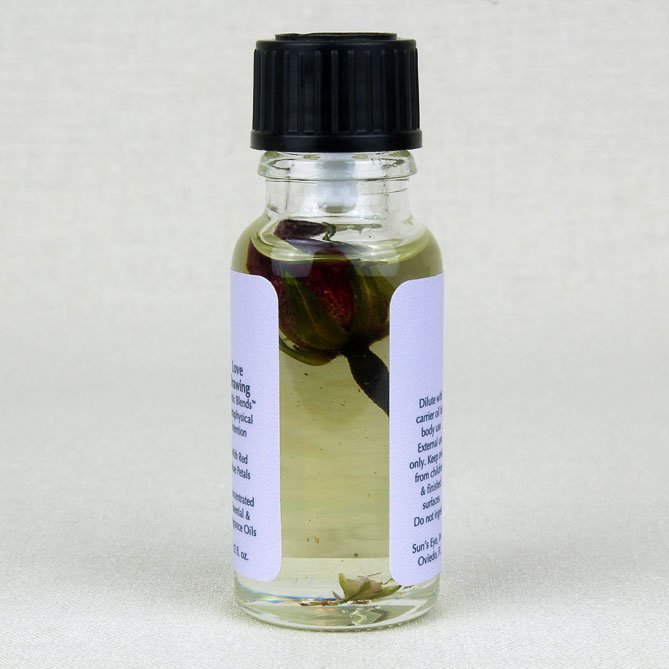 A small clear glass bottle with a black ribbed cap, containing a pale yellow liquid with botanical elements suspended inside. Two white labels are attached to the sides of the bottle, photographed against a neutral light beige textured background.