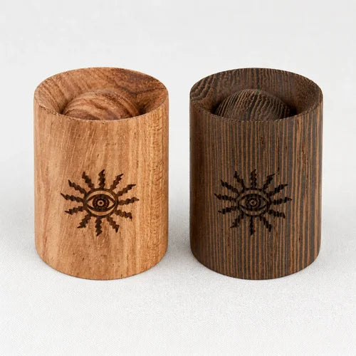Two wooden cylindrical diffusers with engraved eye-in-a-sun symbols, one in light natural wood and one in dark stained wood, shown on a light neutral background.